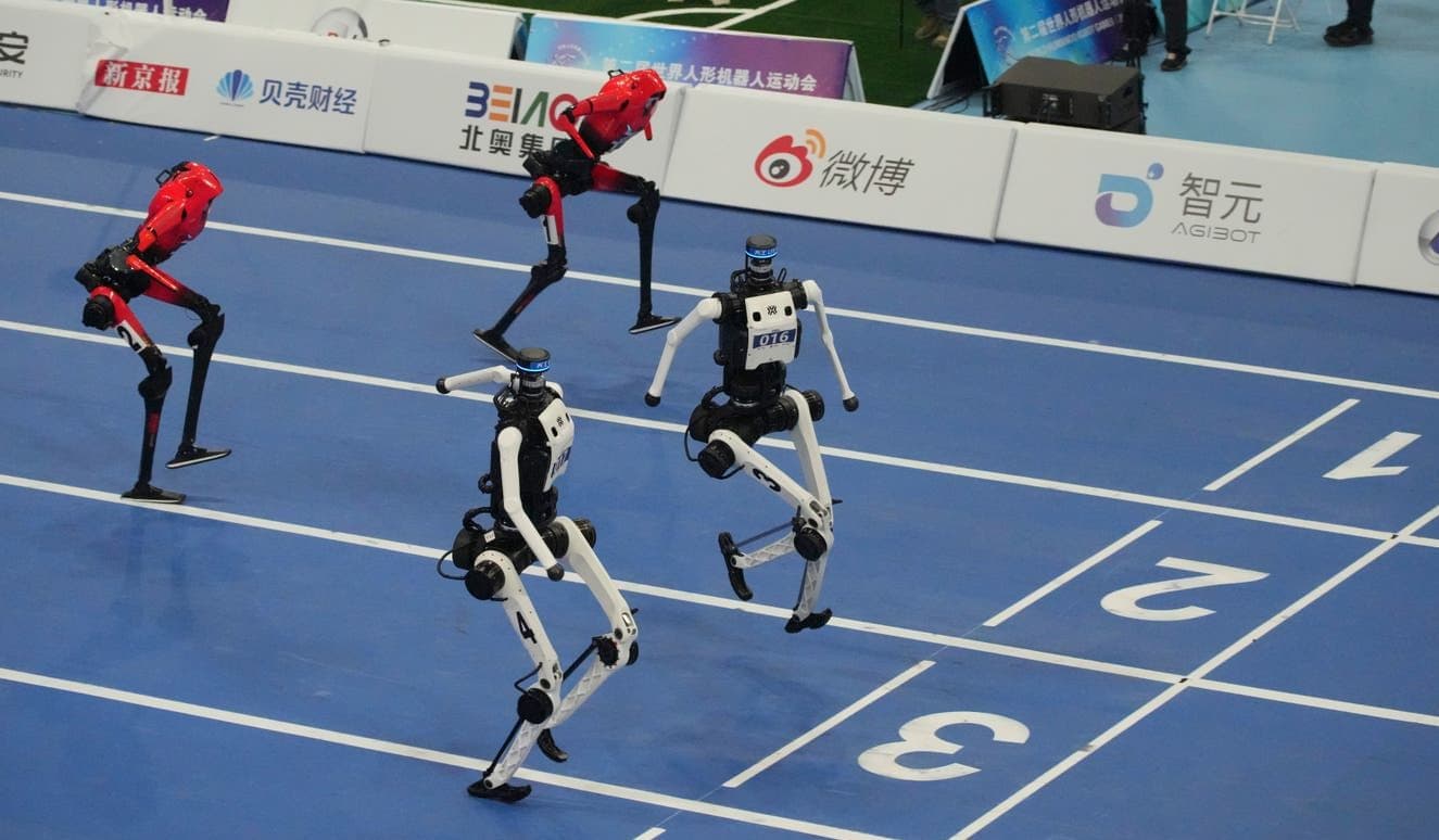 Four bipedal humanoid robots — two red, two black-and-white — mid-stride racing on a blue running track marked with lane numbers, with race sponsor banners in the background.