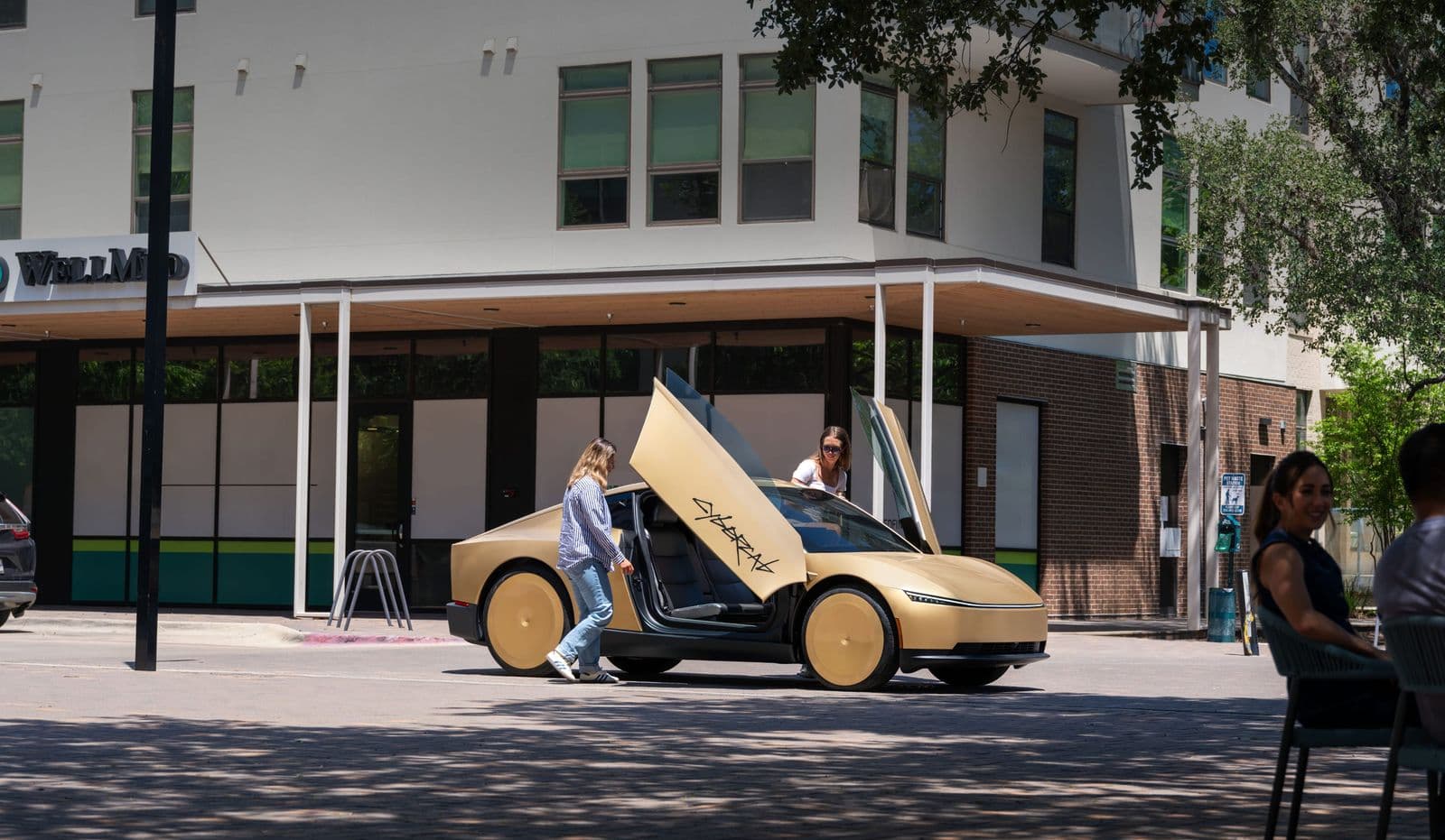 A gold Tesla Cybercab parked on a city street with its scissor-style doors open, as two riders board the vehicle.
