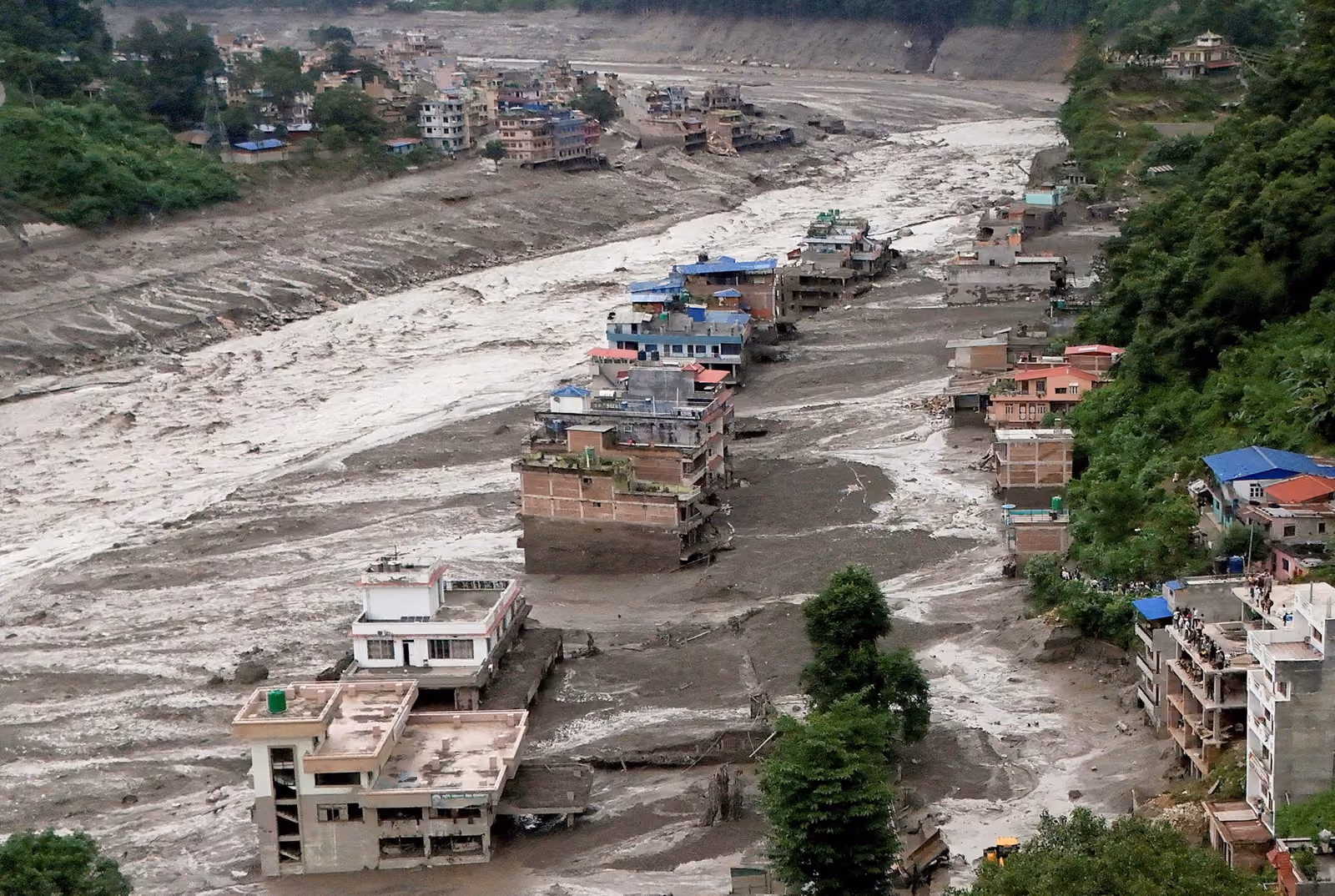Aerial view of a Nepali river valley town devastated by flooding, with multiple buildings standing partially submerged in thick mud and debris carried down by t